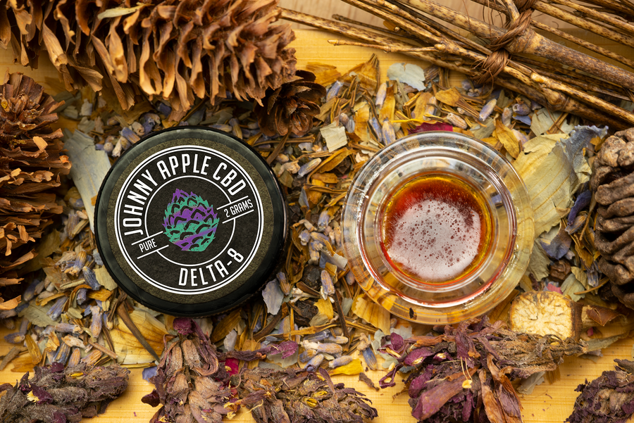 Johnny Apple CBD Delta 8 jar beside an open jar of amber distillate, with pine cones and dried flowers