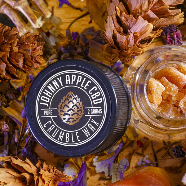 Johnny Apple CBD Crumble Wax jar beside an open jar of crumble wax, with pine cones and dried flowers