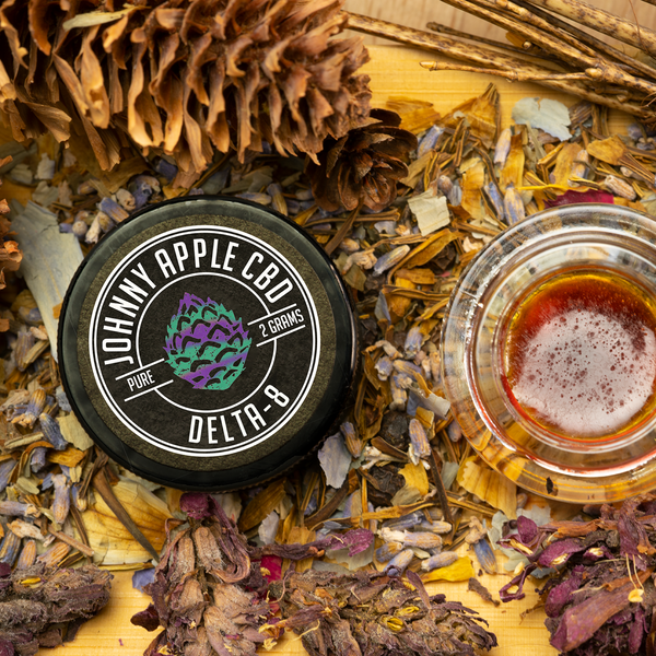Johnny Apple CBD Delta 8 jar beside an open jar of amber distillate, with pine cones and dried flowers
