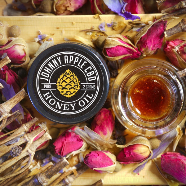 Johnny Apple CBD Honey Oil jar beside an open jar of golden oil, surrounded by dried rose buds