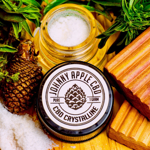 Johnny Apple CBD Pure crystalline jar beside an open jar of crystals, with a pine cone, rosemary and wood blocks